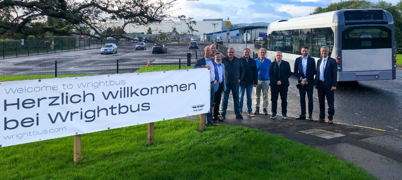 group of men standing at entrance to Wrightbus factory beside a sign that reads welcome to wrightbus in the German language