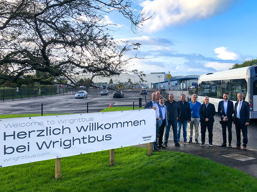group of men standing at entrance to Wrightbus factory beside a sign that reads welcome to wrightbus in the German language