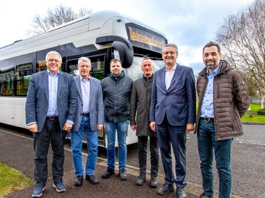 Group of men standing in front of a white single deck, left hand drive hydrogen fuel cell bus