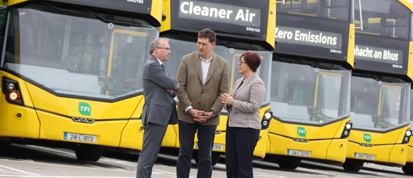 3 people talking in front of a row of parked yellow double deck buses