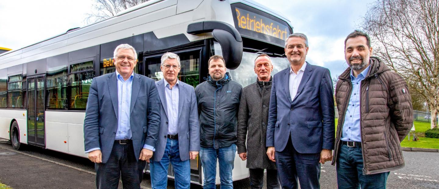 Group of men standing in front of a white single deck hydrogen fuel cell bus. the men are smiling at the camera