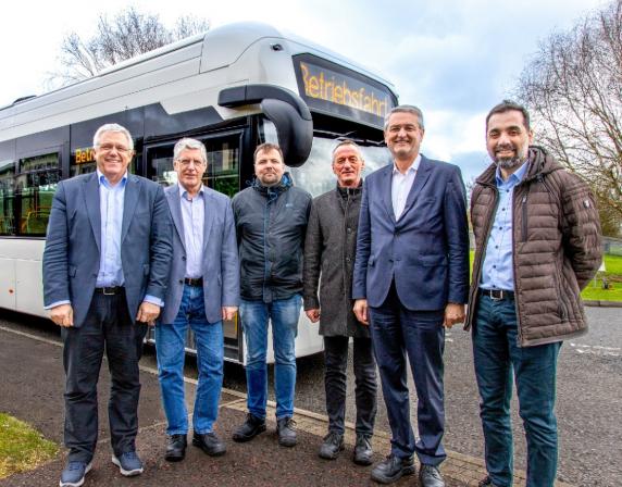 Group of men standing in front of a white single deck hydrogen fuel cell bus. the men are smiling at the camera