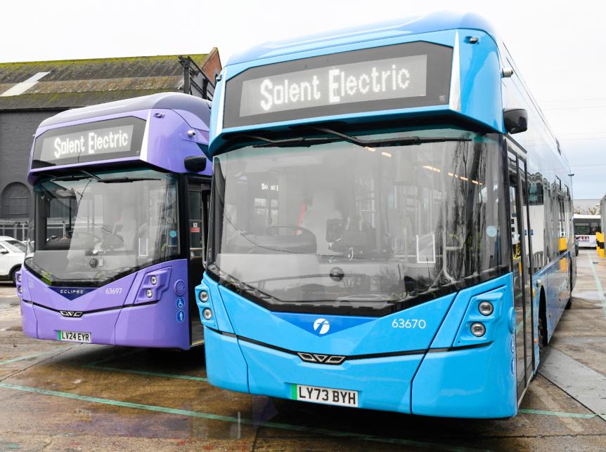 a purple single deck bus and a blue single deck bus parked beside each other in a parking lot