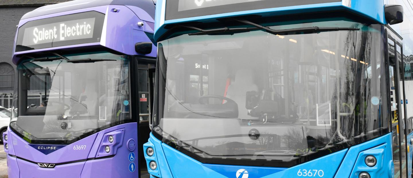 a purple single deck bus and a blue single deck bus parked beside each other in a parking lot