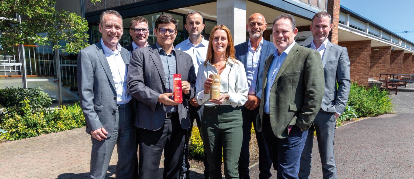 Members of the wrightbus senior team standing outside the factory. A lady in the centre of the photo is holding one award, the man beside her is holding the second award