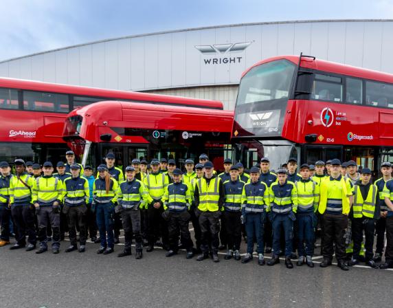 a group of apprentices standing in front of 3 red buses. the wrightbus factory is in the background