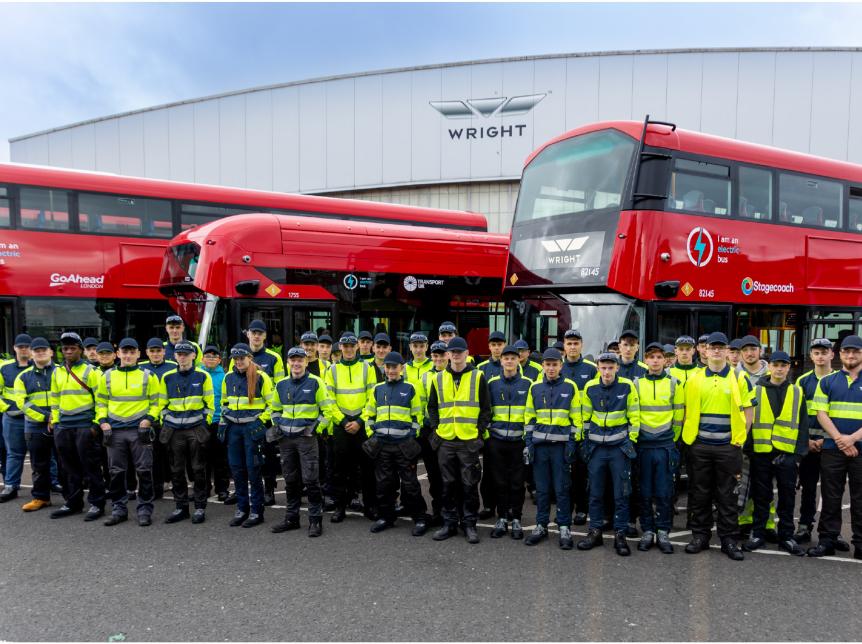 a group of apprentices standing in front of 3 red buses. the wrightbus factory is in the background