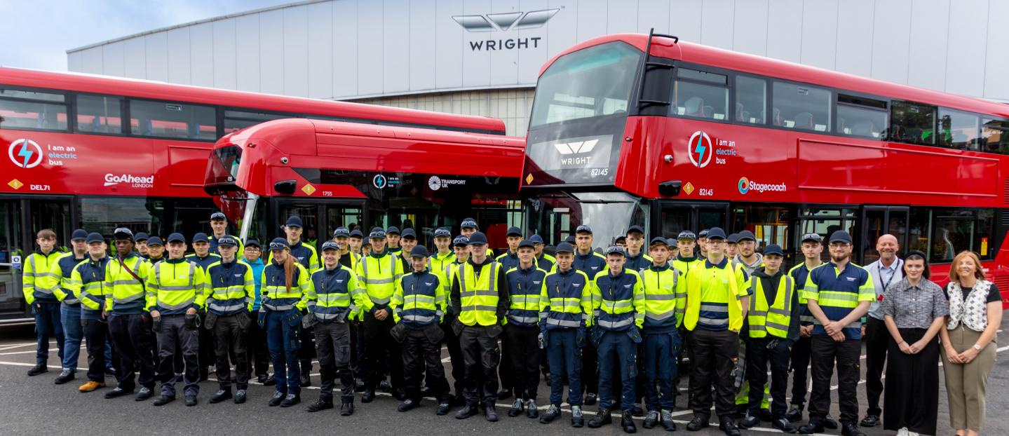a group of apprentices standing in front of 3 red buses. the wrightbus factory is in the background